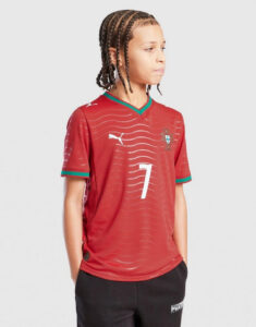 Child wearing a red Portugal soccer jersey with number 7, facing right, with braided hair and hands in pockets.