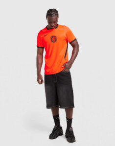 jd_789706_b | UAE SPORT STORE Man in a bright orange soccer jersey with black trim and crest, black shorts, and black sneakers, standing against a light background.