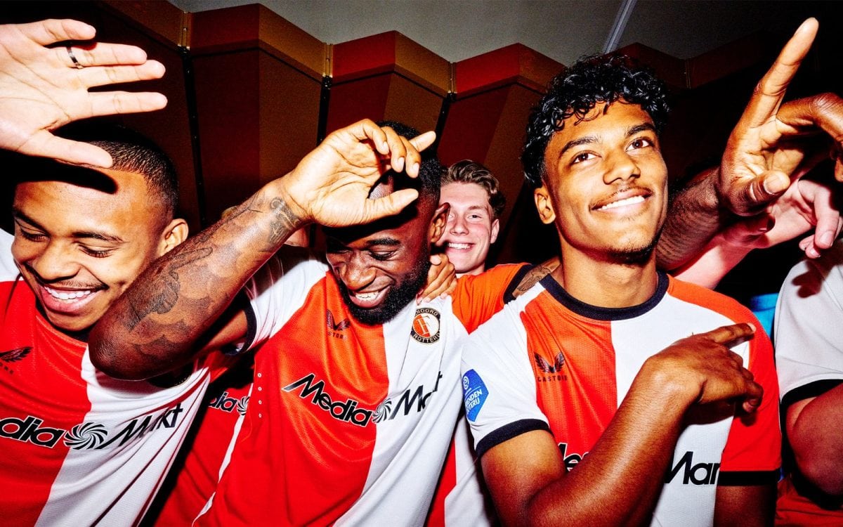 A group of smiling soccer players celebrate, arms around each other in red-and-white striped jerseys.