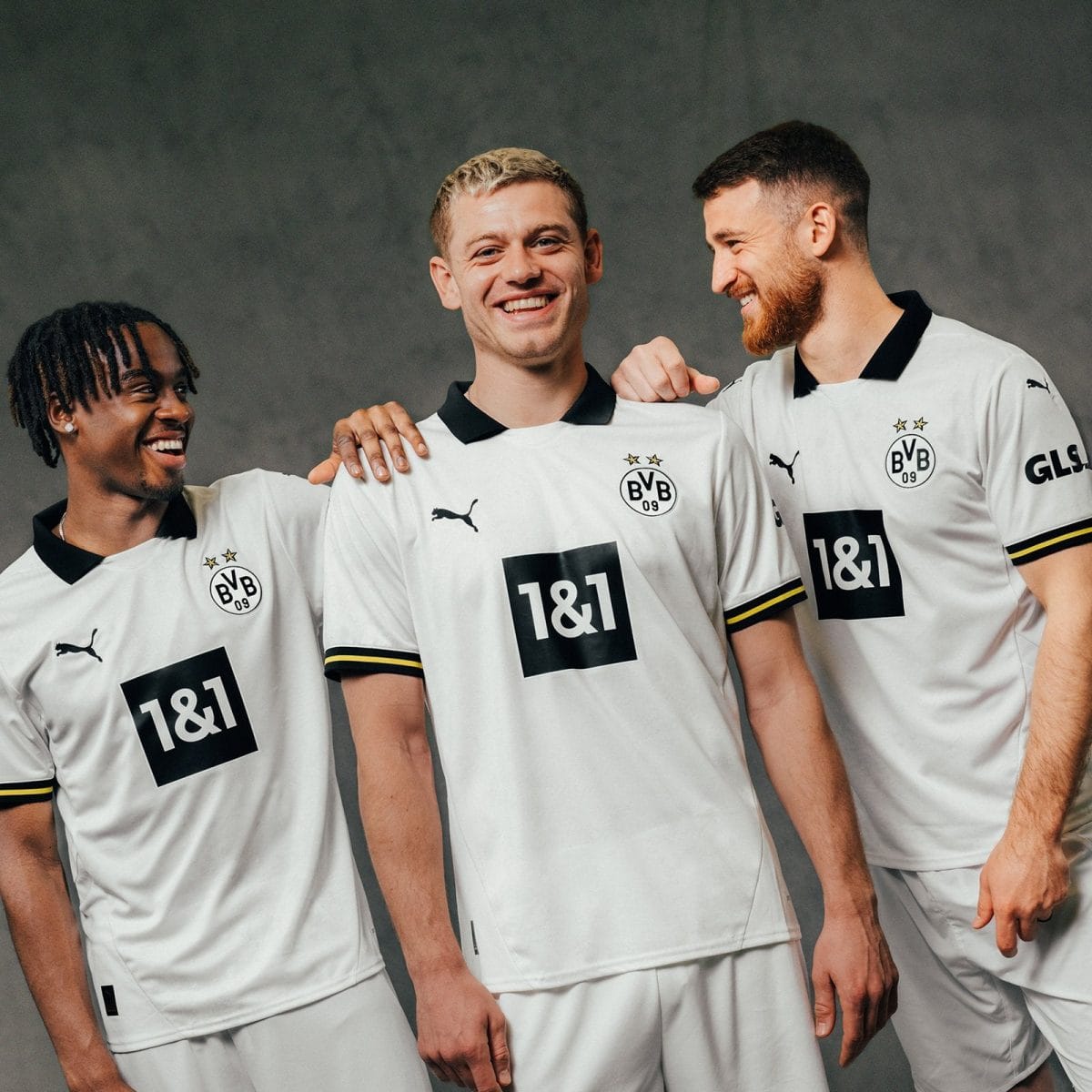 Three male soccer players wearing Borussia Dortmund white jerseys stand together, smiling and engaging with each other in a team photo.