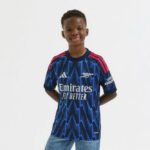 jji9550_f | UAE SPORT STORE Smiling boy wearing a blue Arsenal football jersey with red accents and the 'Emirates Fly Better' logo, standing with hands behind his back.