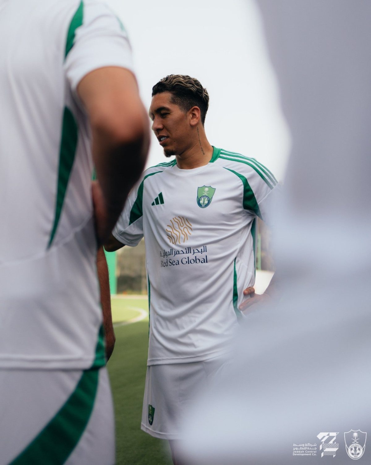 Soccer player in a white and green kit talking with teammates during a huddle on the field, sponsor logo visible on the jersey.