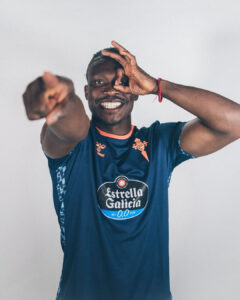 Smiling man in a blue sports jersey makes an OK gesture around his eye and points at the camera playfully.