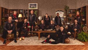 A group of twelve people in matching jackets pose in a retro living room with shelves, a chessboard on the coffee table, and plant decor for a team photo.