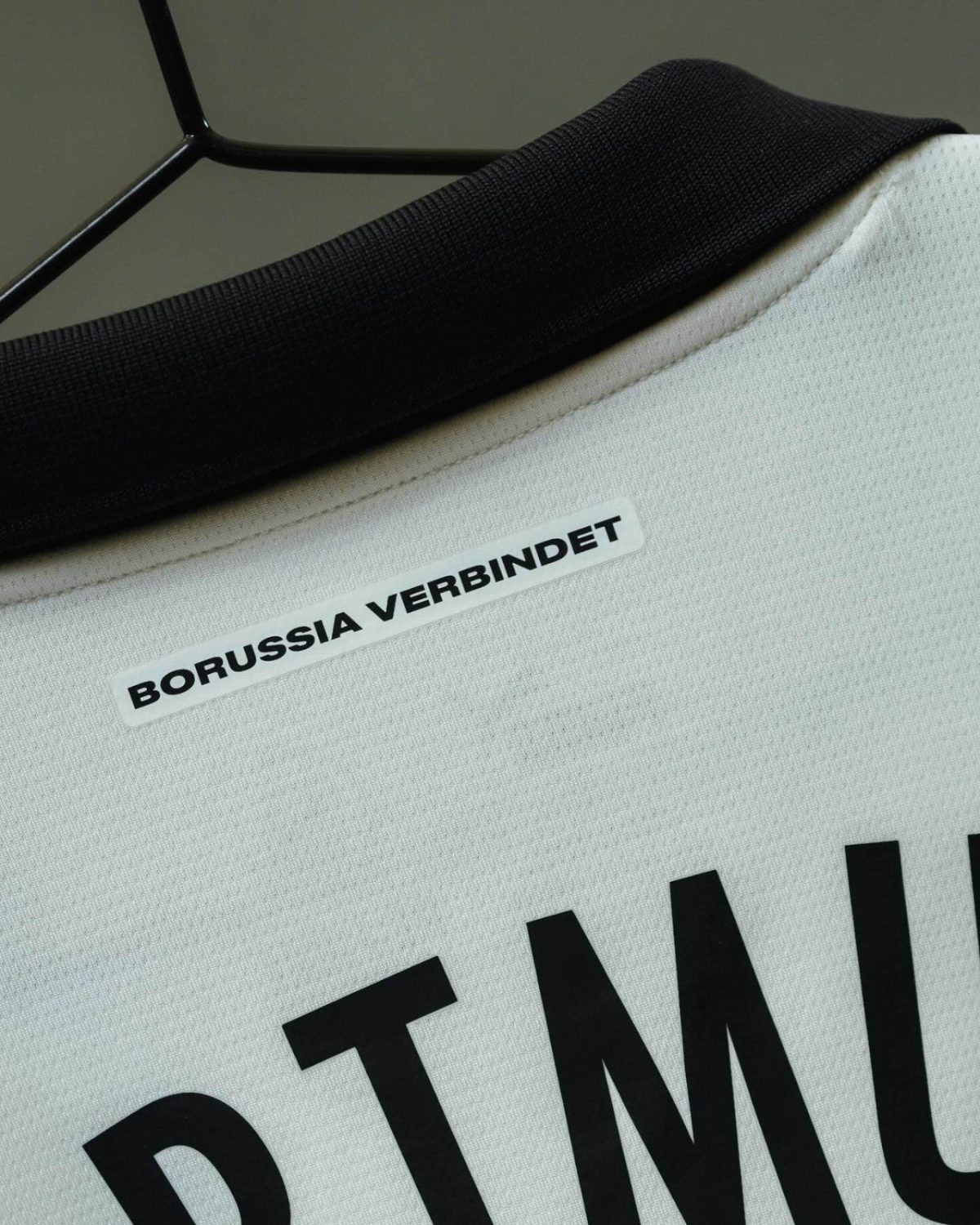 Close-up of a white sports jersey with a black collar on a hanger, showing a white label that reads 'BORUSSIA VERBUNDET'