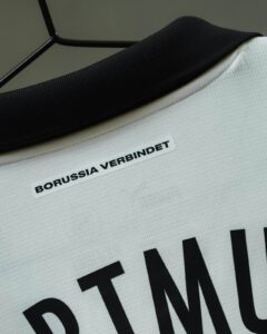 Close-up of a white sports jersey with a black collar on a hanger, showing a white label that reads 'BORUSSIA VERBUNDET'
