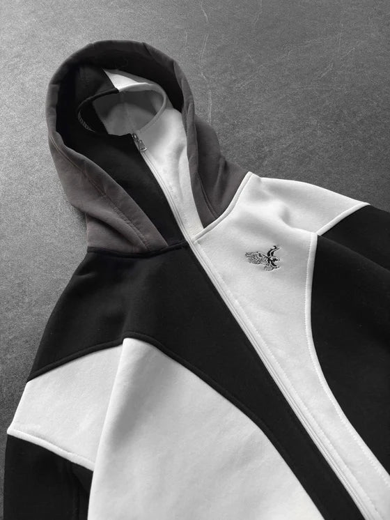 Color-block zip hoodie in black, white, and gray with a diagonal panel design and a small embroidered eagle logo on the chest.