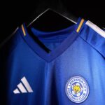 Blue Leicester City football jersey on a hanger, close-up of the club crest and Adidas logo on the chest area.