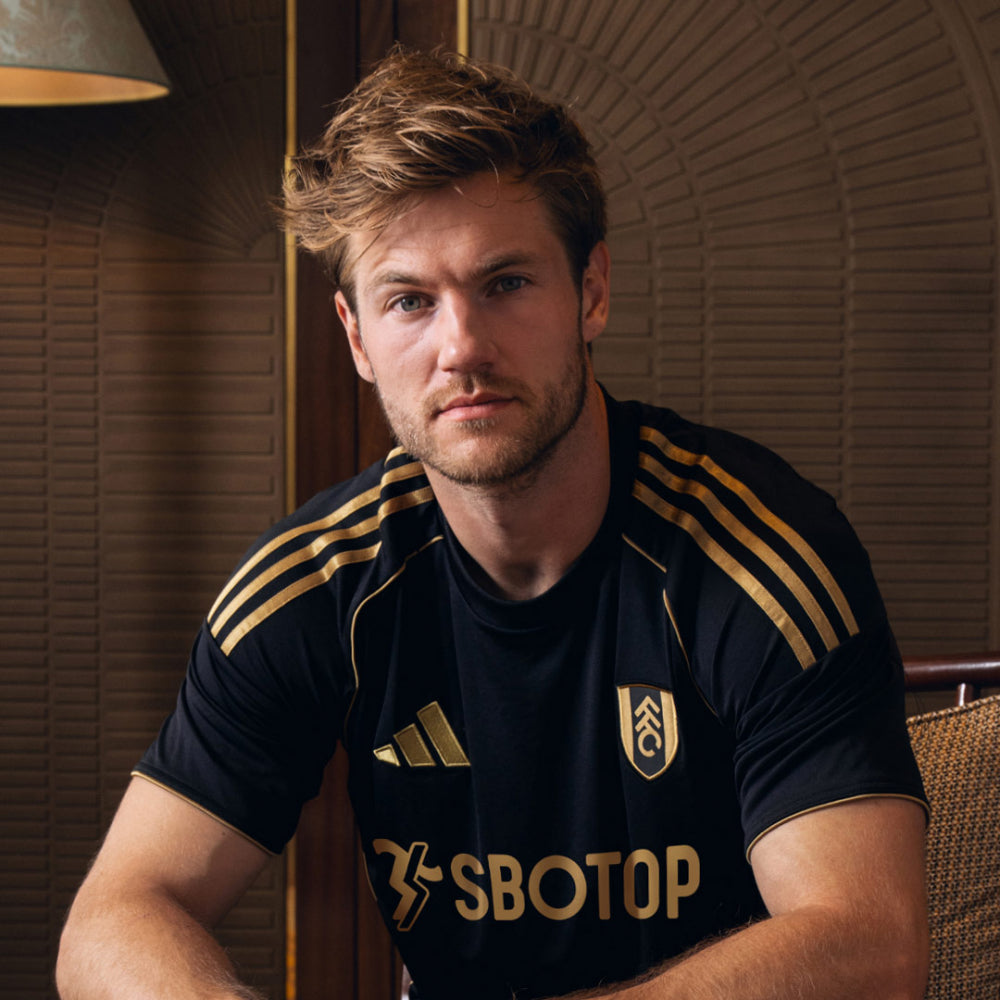 ko | UAE SPORT STORE Portrait of a man with short blond hair wearing a black soccer jersey with gold stripes and a crest, looking at the camera indoors.