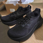 Black running shoes with chunky soles sit on a wooden table, with an open shoebox in the background.