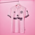 Pink Brentford FC football jersey on a hanger against a pink background, featuring Hollywood Bets sponsor and club crest such as the Brentford badge and Umbro logo.