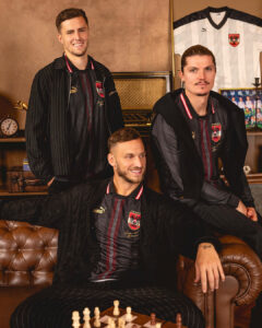 Three men in matching black pinstripe soccer jerseys pose in a cozy room with a chessboard and vintage decor behind them, looking at the camera.