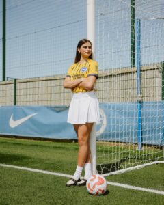 Young woman in a yellow soccer jersey and white skirt stands with arms crossed beside a goal post, a soccer ball at her feet on a sunny field.