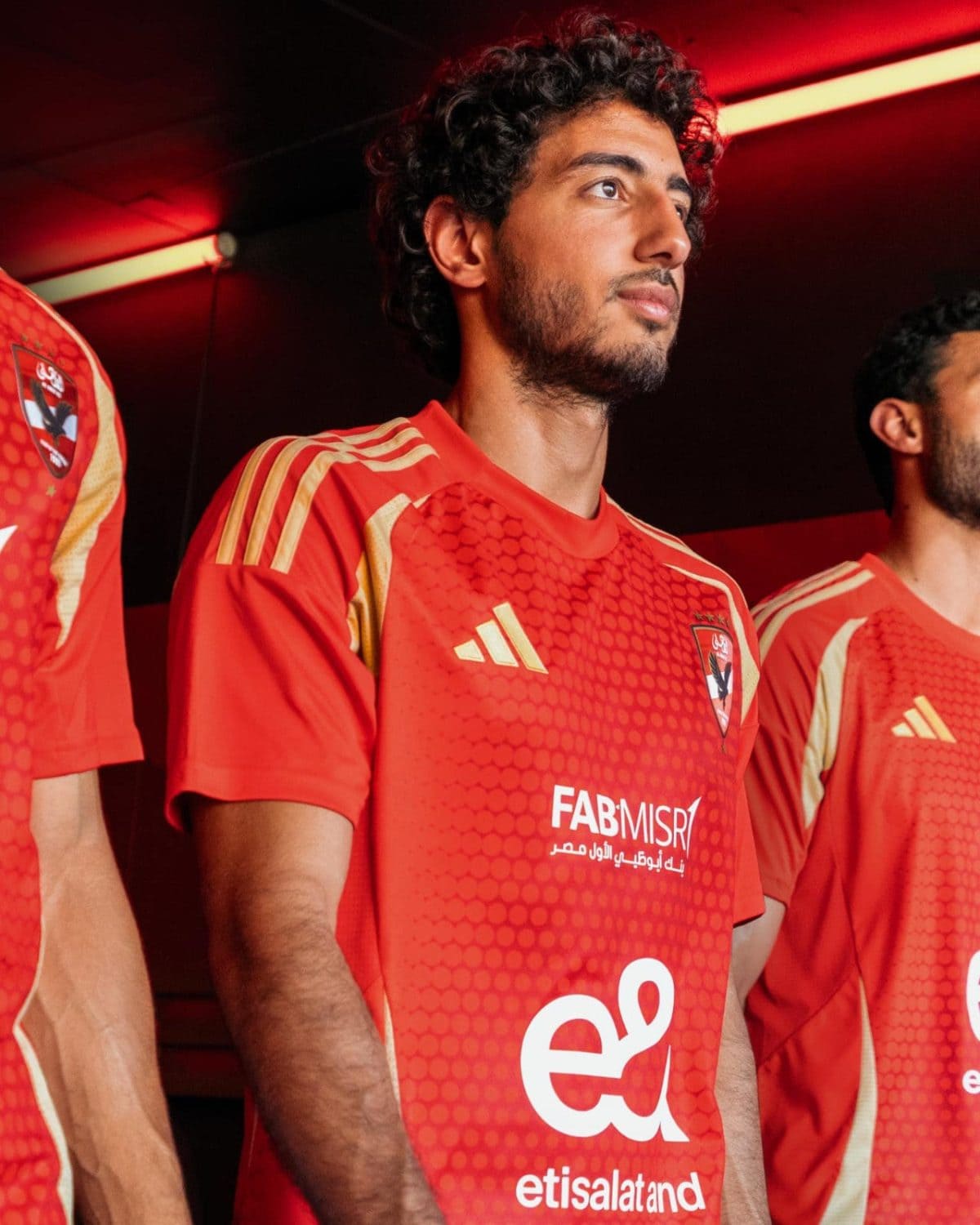Young man in a red athletic jersey standing with teammates, looking upward in an indoor setting with red lighting.