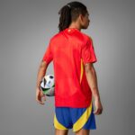 Person seen from behind in a red soccer jersey with yellow accents, holding a soccer ball at their left hip; wearing blue and yellow shorts, gray background