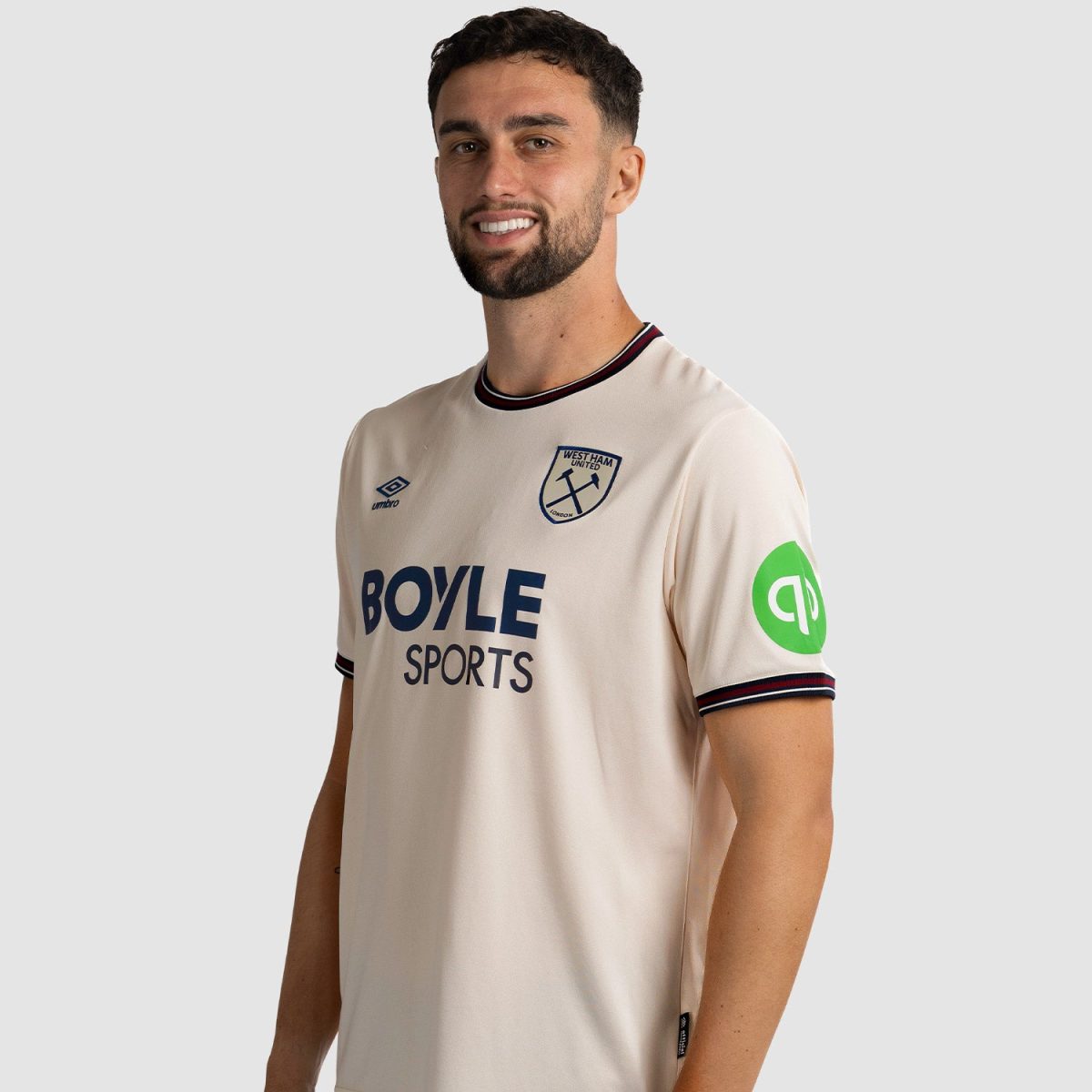 Male model wearing a beige West Ham United jersey with Boyle Sports sponsor, Umbro logo, and a green patch on the sleeve; smiling.