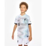 Young boy in a white Liverpool FC youth jersey with blue swirl pattern and Standard Chartered logo, standing against a white background.