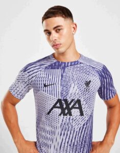 Male model wearing a purple and white patterned soccer jersey with the AXA sponsor and Liverpool FC crest on the chest, standing with hands on hips against a light background.