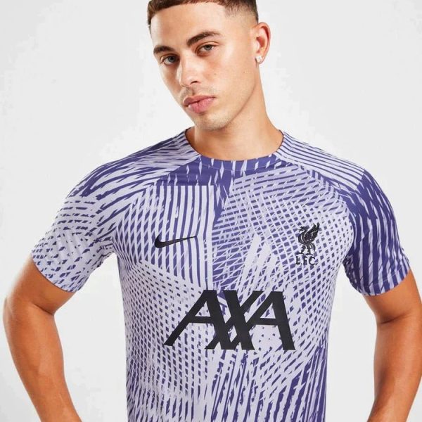 Male model wearing a purple and white patterned soccer jersey with the AXA sponsor and Liverpool FC crest on the chest, standing with hands on hips against a light background.