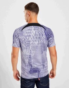 Back view of a man wearing a purple abstract-patterned T-shirt, standing against a light background, with dark jeans.