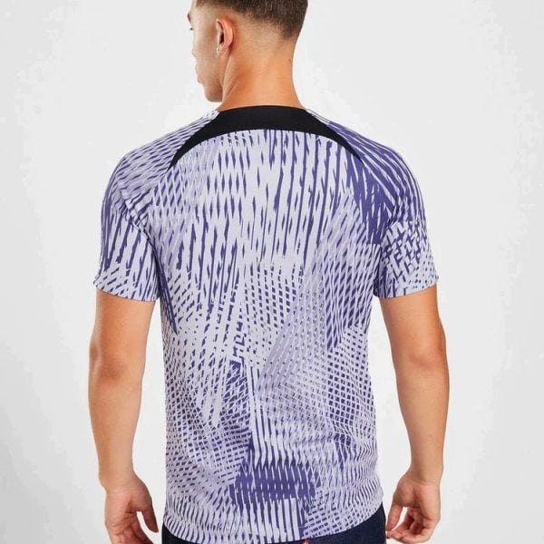 Back view of a man wearing a purple abstract-patterned T-shirt, standing against a light background, with dark jeans.