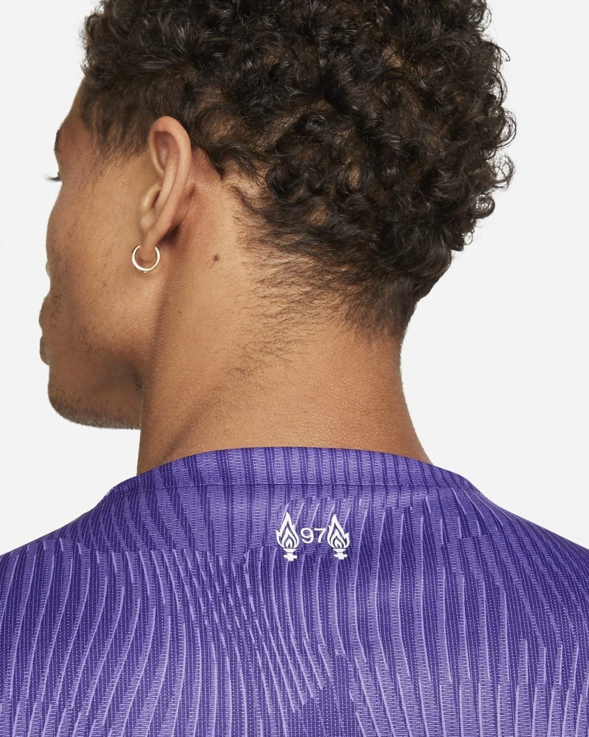 Back view of a person with short, curly hair and a small gold hoop earring, wearing a purple striped shirt with the number 97 and flame icons on the back.
