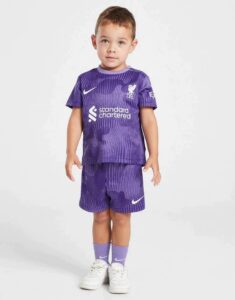 Toddler wearing a purple Liverpool FC soccer kit (jersey and shorts) with white sneakers, standing against a light background.
