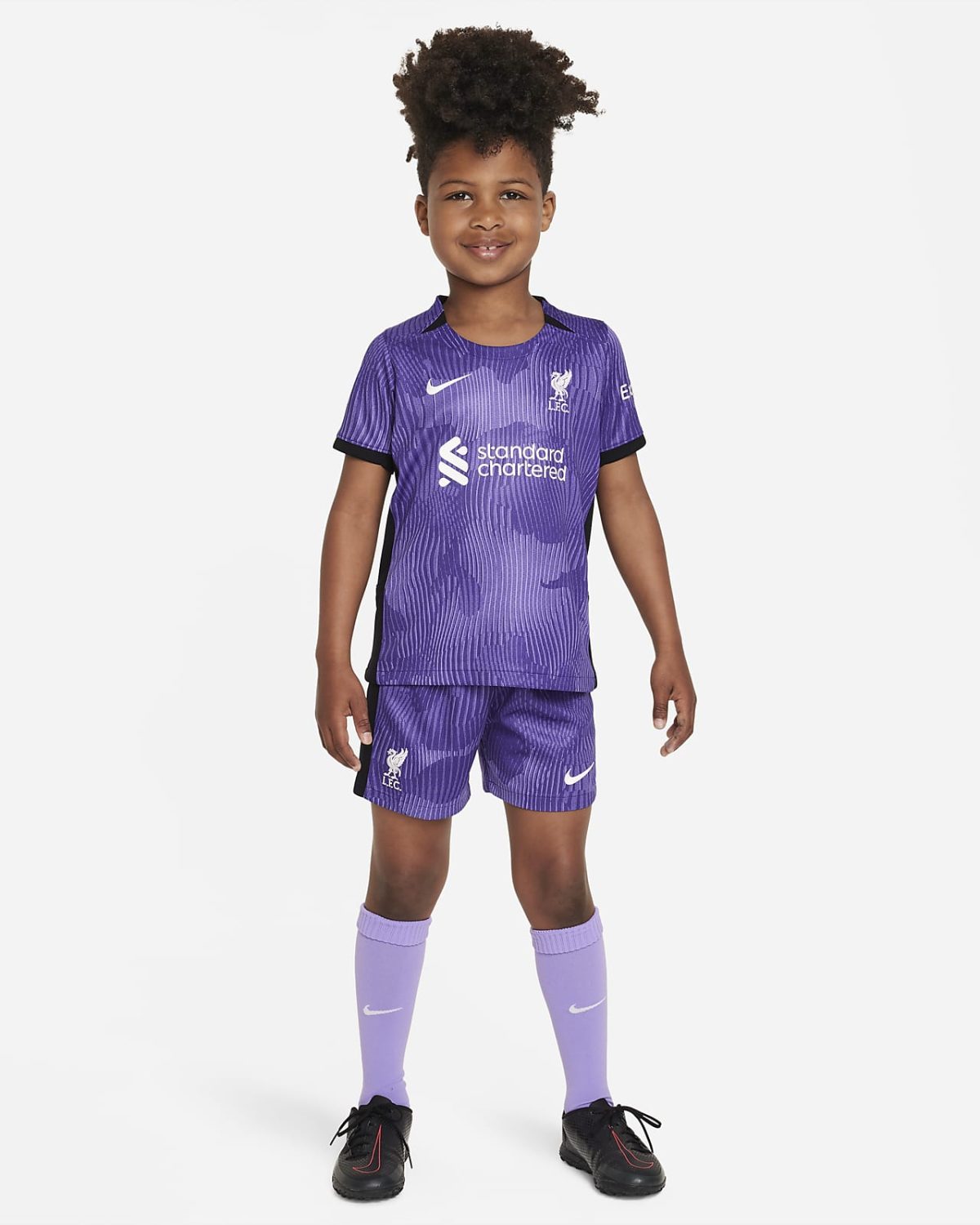 Child wearing a purple Liverpool FC soccer kit (jersey and shorts) and lavender socks, standing and smiling at camera.