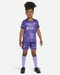 Child wearing a purple Liverpool FC soccer kit (jersey and shorts) and lavender socks, standing and smiling at camera.