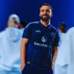 Man wearing a dark blue Manchester United jersey with Snapdragon logo, standing with hands in pockets and smiling, blurred people in white hoodies behind him.