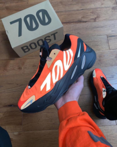 Bright orange and black Yeezy Boost 700 sneaker held in hand on a wooden floor, with a '700 Boost' box in the background.