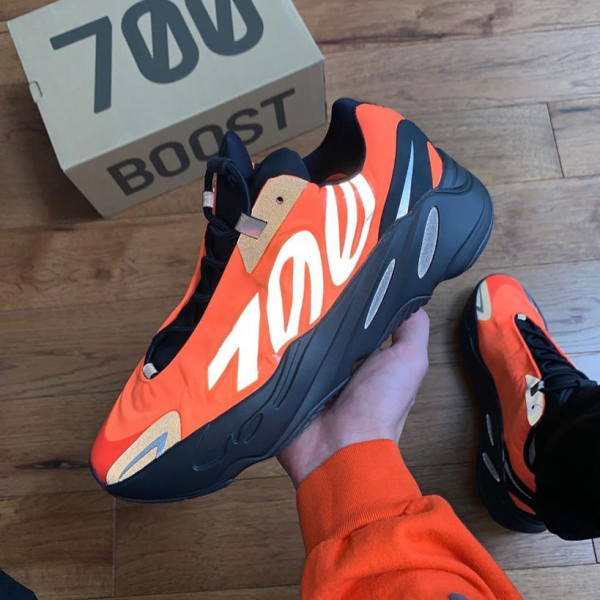 Bright orange and black Yeezy Boost 700 sneaker held in hand on a wooden floor, with a '700 Boost' box in the background.