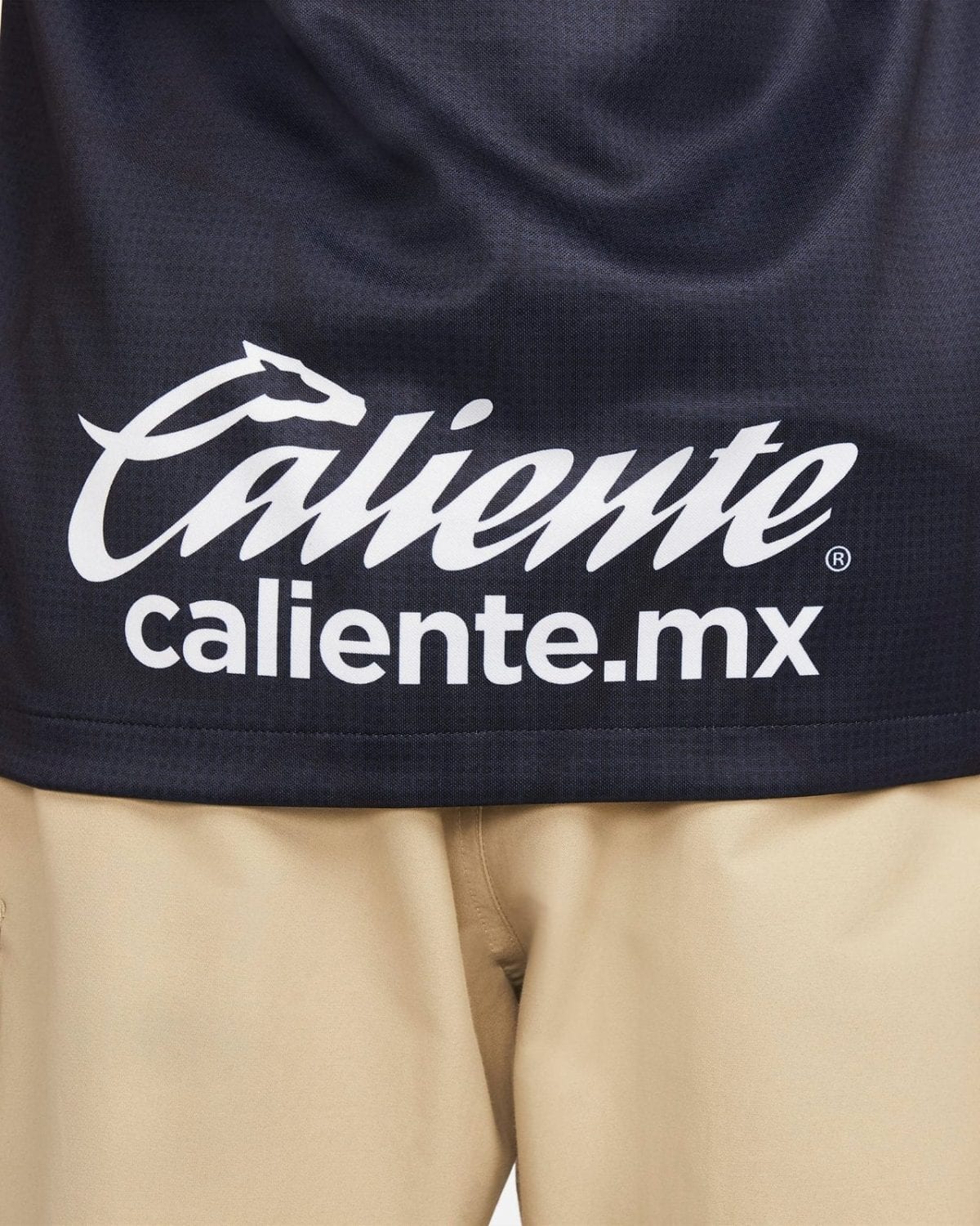 Navy fabric with white 'Caliente' script and 'caliente.mx' text branding at the bottom center on a dark shirt surface.