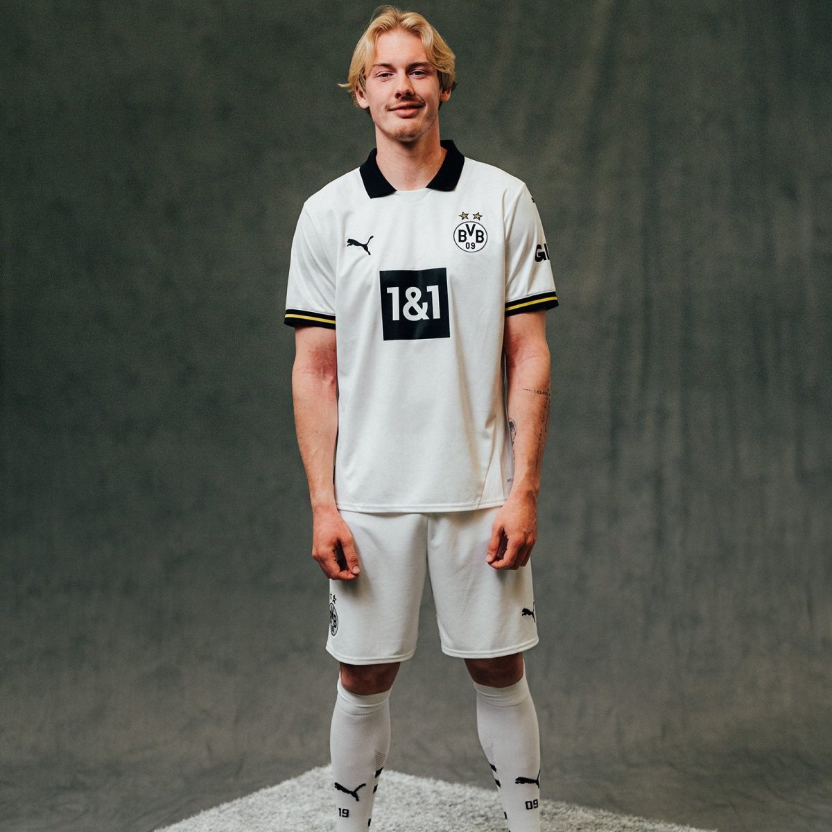 Young male footballer in a white Borussia Dortmund kit stands facing the camera against a dark gray backdrop, hands at his sides.