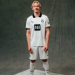 Young male footballer in a white Borussia Dortmund kit stands facing the camera against a dark gray backdrop, hands at his sides.