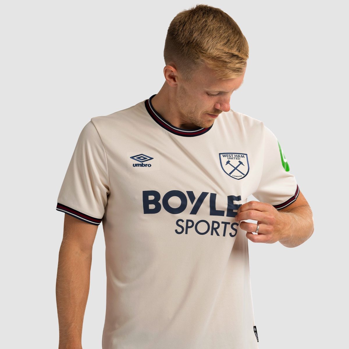 Man wearing a cream West Ham United jersey with Umbro logo and Boyle Sports sponsor, looking down at his chest as he adjusts the shirt.