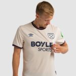 Man wearing a cream West Ham United jersey with Umbro logo and Boyle Sports sponsor, looking down at his chest as he adjusts the shirt.