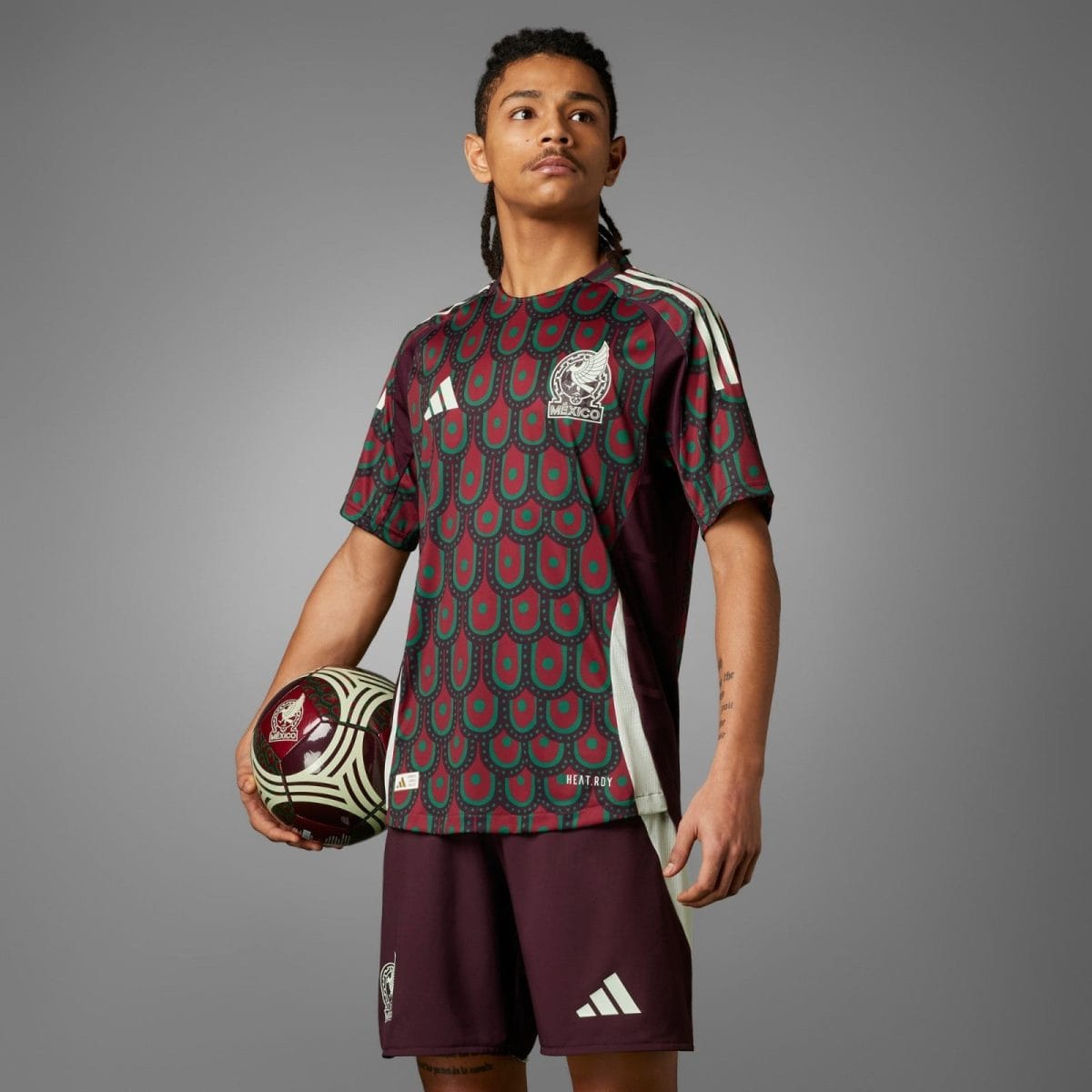 Male soccer player wearing a maroon and teal patterned jersey with white Adidas stripes, holding a patterned soccer ball against a gray background, crest visible on chest
