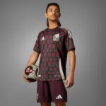 lysfKch8ylx0LmC | UAE SPORT STORE Male soccer player wearing a maroon and teal patterned jersey with white Adidas stripes, holding a patterned soccer ball against a gray background, crest visible on chest