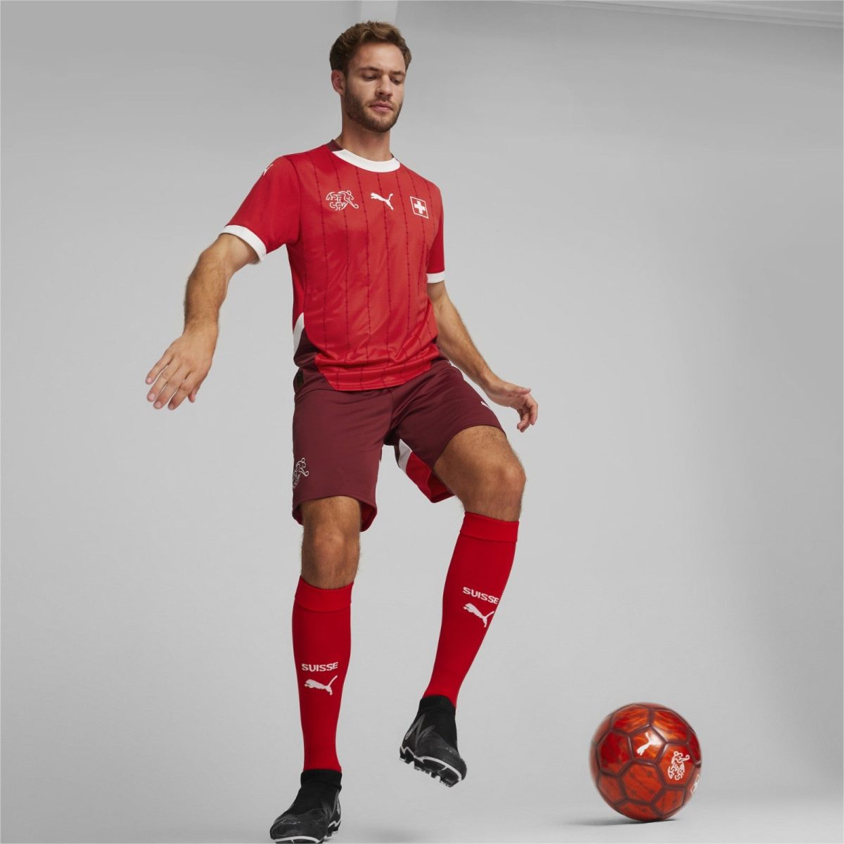 Male soccer player in a red Swiss team kit, mid-action kicking an orange soccer ball in a neutral studio setting.