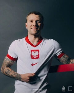 Polish footballer wearing a white jersey with red eagle crest and Nike logo, smiling with tattoos on both arms against a dark background.