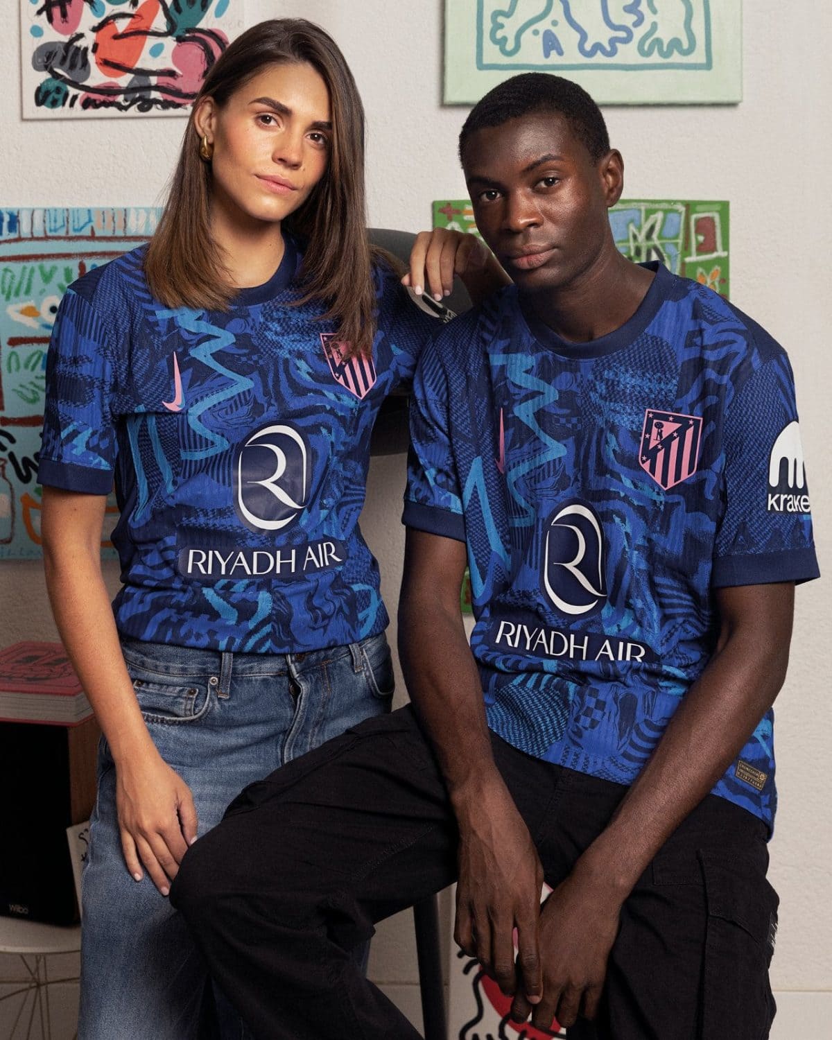 Two young adults sit side by side in blue Riyadh Air soccer jerseys with abstract patterns; a woman rests a hand on a man’s shoulder in front of colorful wall art.