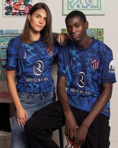 mGeV9NIr5Qg9vAG | UAE SPORT STORE Two young adults sit side by side in blue Riyadh Air soccer jerseys with abstract patterns; a woman rests a hand on a man’s shoulder in front of colorful wall art.