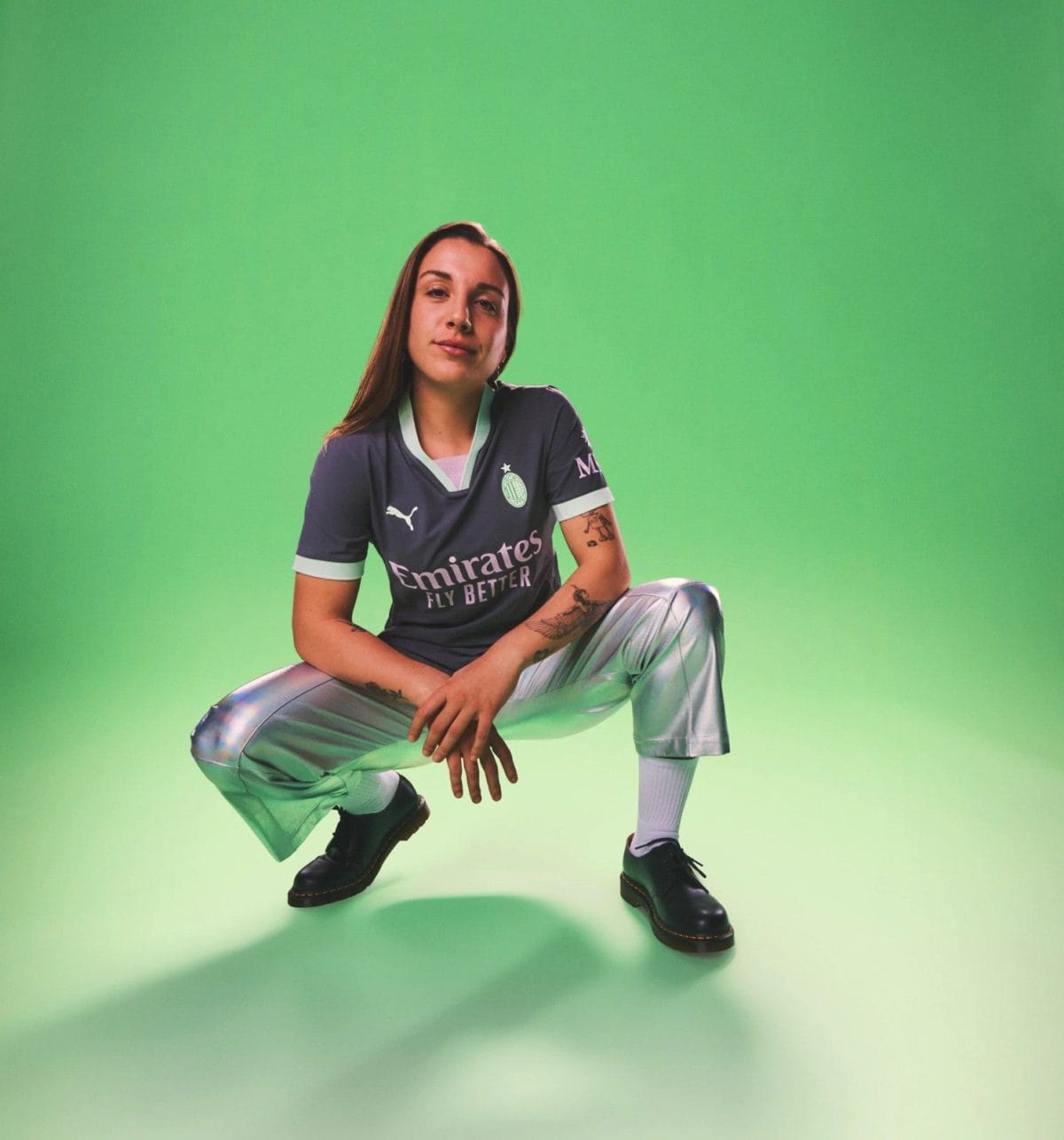 Person crouching against a bright green backdrop, wearing a dark jersey with 'Emirates FLY BETTER' and light-colored pants; tattoos visible on the arm.