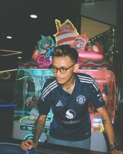 Man wearing a dark Manchester United jersey and glasses leans on an arcade table in a neon-lit arcade with colorful cartoon decorations behind him.