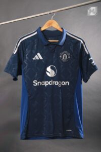 Navy Manchester United football jersey on a wooden hanger, featuring Snapdragon sponsor logo on the front and white club crest on the chest.