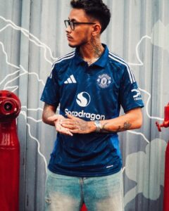 Portrait of a tattooed man wearing a blue Manchester United jersey with glasses, standing outdoors against a patterned wall.
