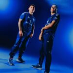 Two men in blue athletic uniforms posing together in a blue-lit studio? with confident stances and hands on hips.