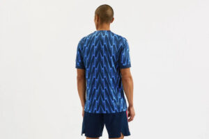 Rear view of a man wearing a blue zigzag-patterned t-shirt and dark shorts, standing in a neutral space.