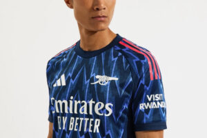 Man wearing a blue Arsenal football jersey with Emirates Fly Better sponsor and Visit Rwanda on the sleeve, close-up of upper body.